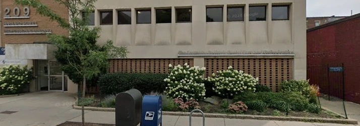 A street view of a two‑story building with rectangular windows and landscaped bushes