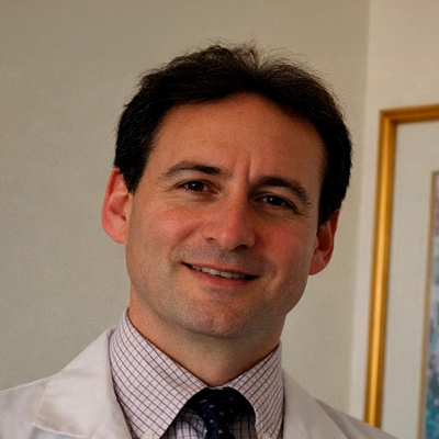 Dr. Jeffrey Bernstein wearing a white lab coat and a patterned dress shirt with a tie, standing indoors near a framed artwork